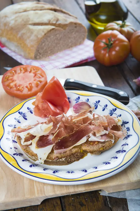 Zwei Scheiben geröstetes Bauernbrot mit Tomate und Serranoschinken auf einem blau gemusterten Teller, daneben eine aufgeschnittene Tomate.