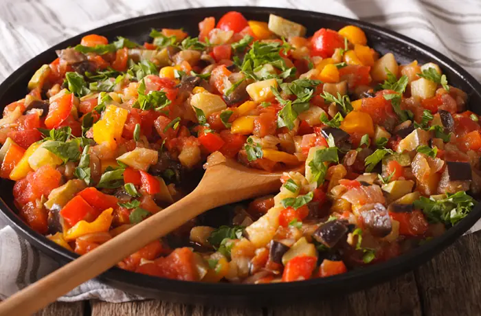 Eine Pfanne mit geschmortem Gemüse aus Paprika, Zucchini, Auberginen und Tomaten, dazu ein Holzlöffel.