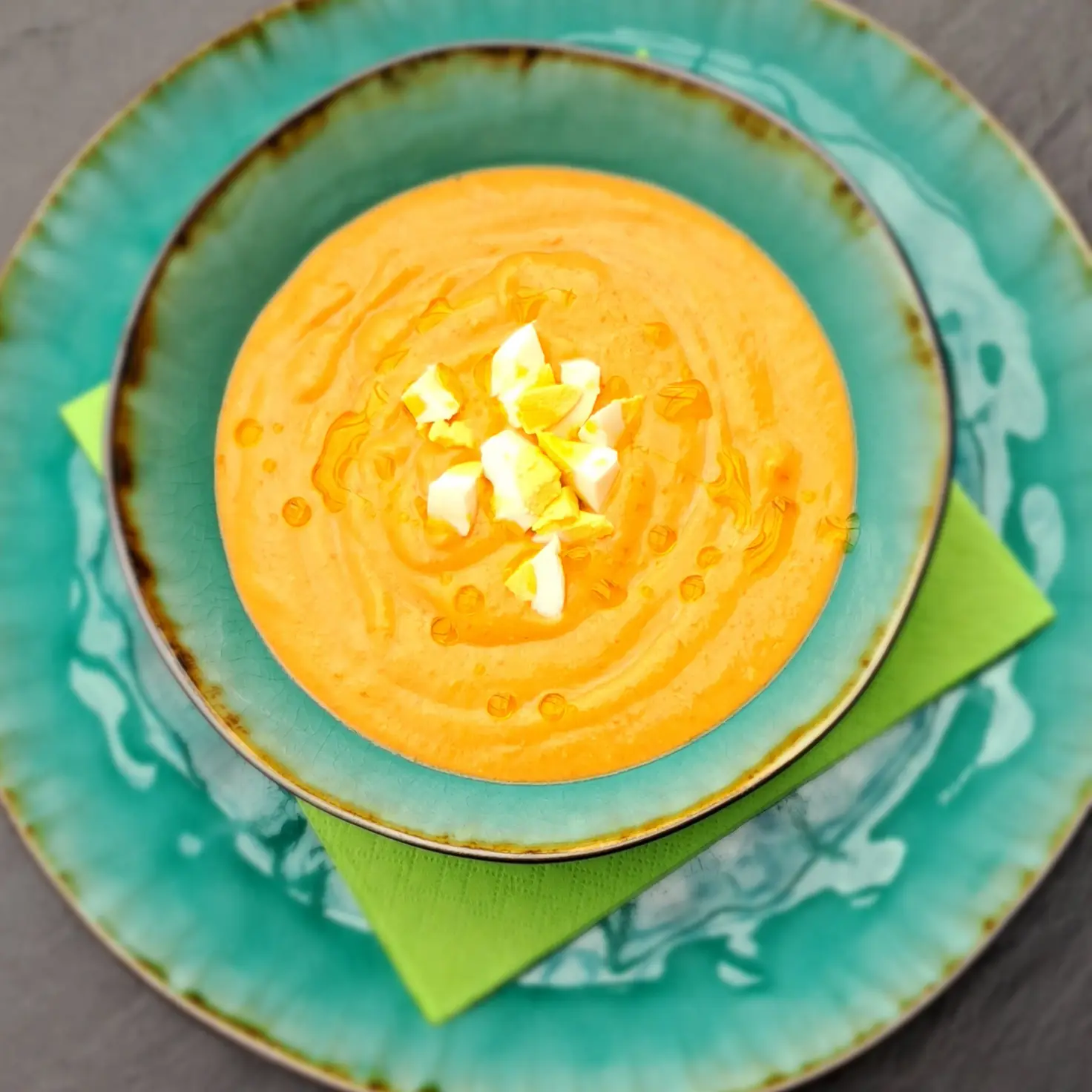 Eine Schale kalte Salmorejo auf einem türkisen Teller, mit hartgekochtem Ei und einem Schuss Olivenöl bestreut.